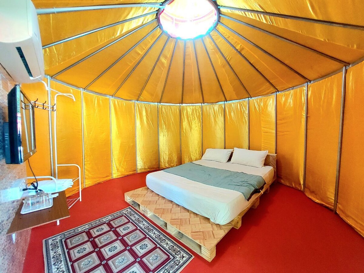 An airy yurt features a spacious bed positioned on a wooden platform, surrounded by golden fabric walls. A flat-screen television is mounted nearby, and a small table is set against one wall. The room is illuminated by natural light from a central circular window.