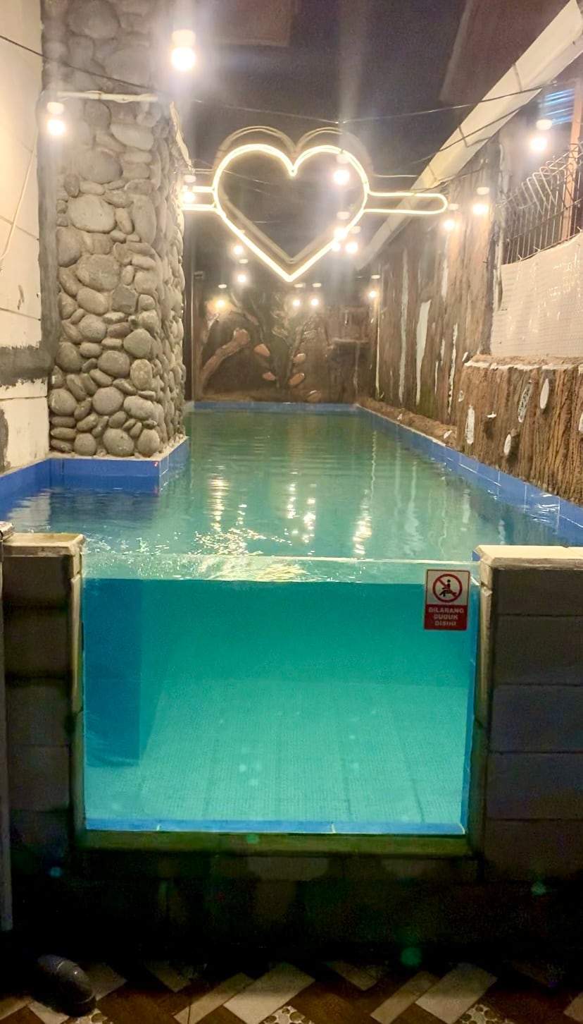 A private swimming pool is showcased, with shimmering blue water reflecting soft lighting. Stone walls provide a natural backdrop, enhanced by decorative fixtures, including a heart-shaped light above the pool area. The tranquil setting is designed for relaxation.