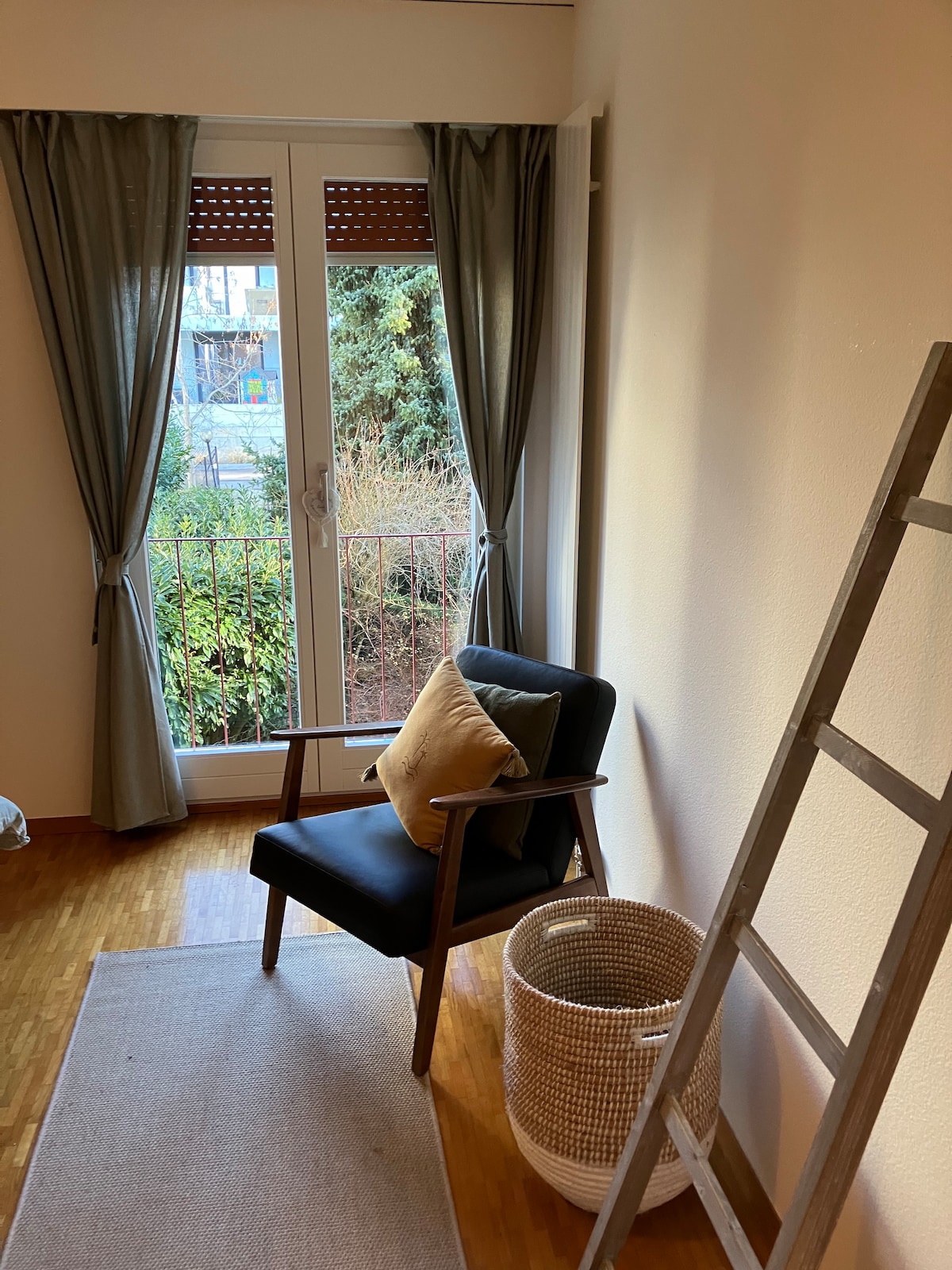 A corner of the room features a comfortable chair with a decorative pillow, positioned beside a small balcony door. Natural light filters through sheer curtains, illuminating a simple rug and a woven basket nearby. A wooden ladder is placed against the wall.