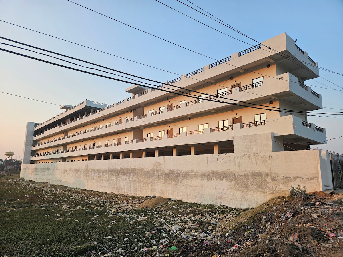 A modern, multi-story building is depicted, featuring a clean façade and multiple balconies. The structure is set against an open sky, with power lines overhead. The surrounding area includes grassy patches, hinting at ongoing development.