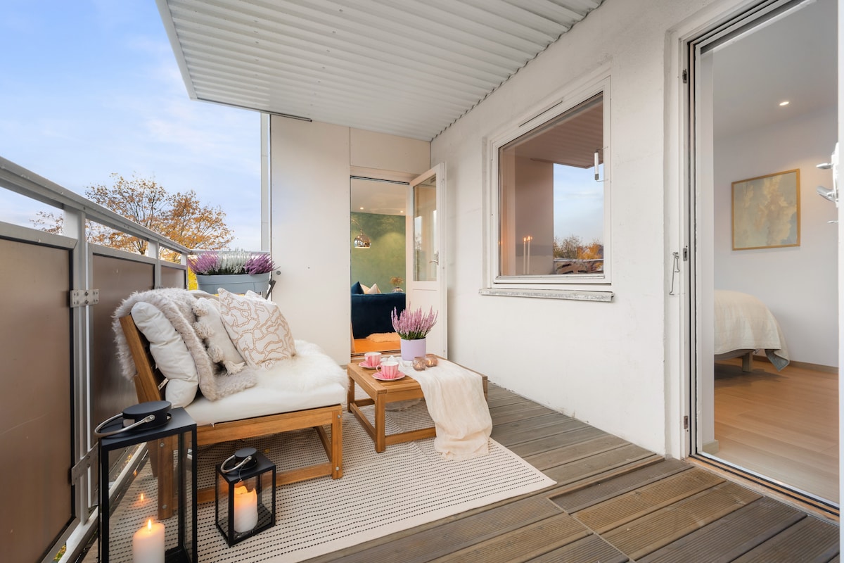 A cozy balcony space is presented with a wooden seating arrangement featuring plush cushions. A small table is set with snacks and decorative items. Light-colored outdoor flooring complements the soft area rug, while a candle adds warmth. Nearby, an open window connects to the interior.