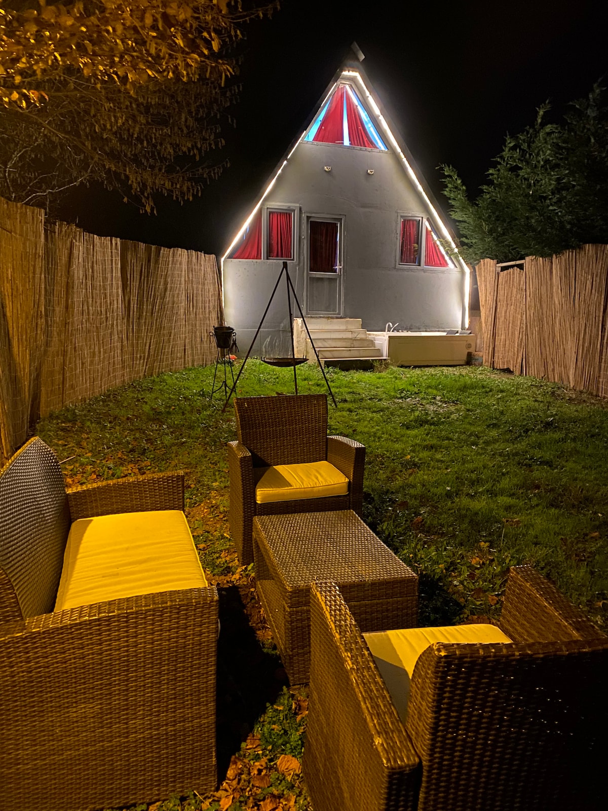 A cozy outdoor seating area is highlighted by wicker furniture with yellow cushions. Soft lighting outlines a triangular-shaped house, while a well-kept grassy yard provides a tranquil backdrop. The lively atmosphere is enhanced by the privacy offered by tall wooden fencing.