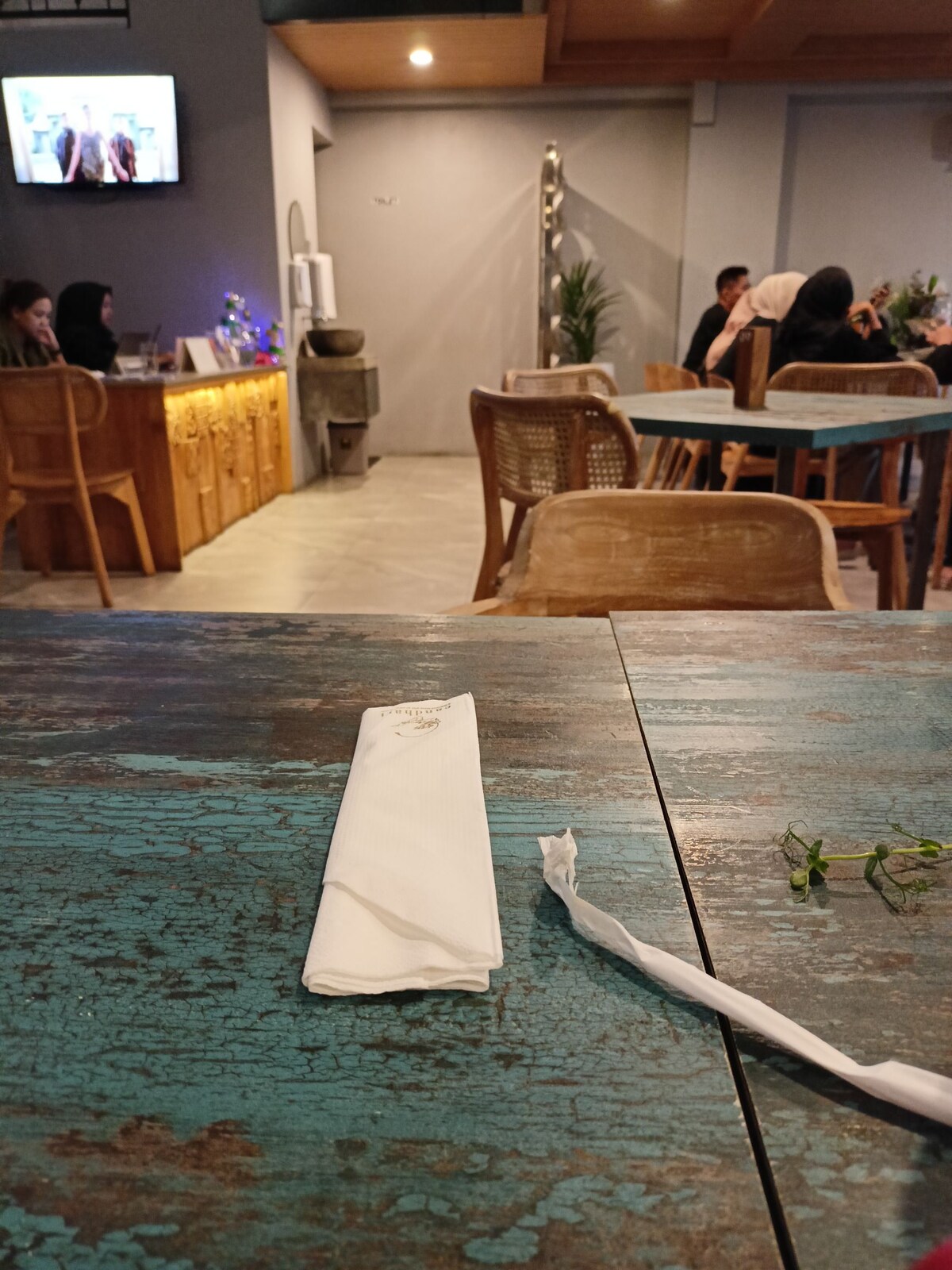A rustic wooden table is set with a folded napkin and a small piece of greenery. In the background, a softly lit seating area is visible, with several patrons enjoying their time. A television screen can be seen, adding to the cozy ambiance.
