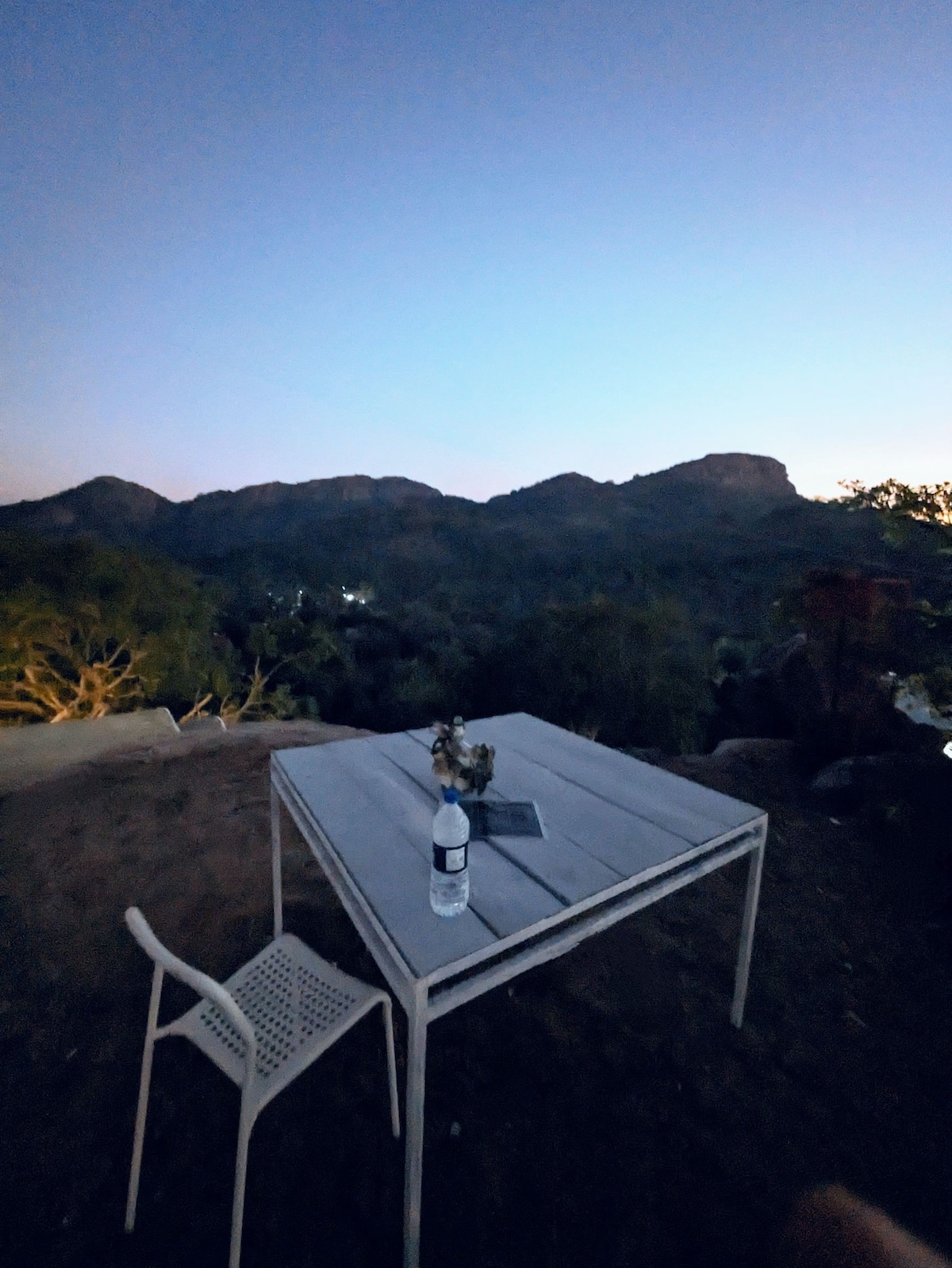 An outdoor table sits on a hillside, positioned to capture views of the surrounding mountains at dusk. A single chair accompanies the table, which holds a water bottle and a small vase with greenery. The early evening sky presents soft hues transitioning into night.