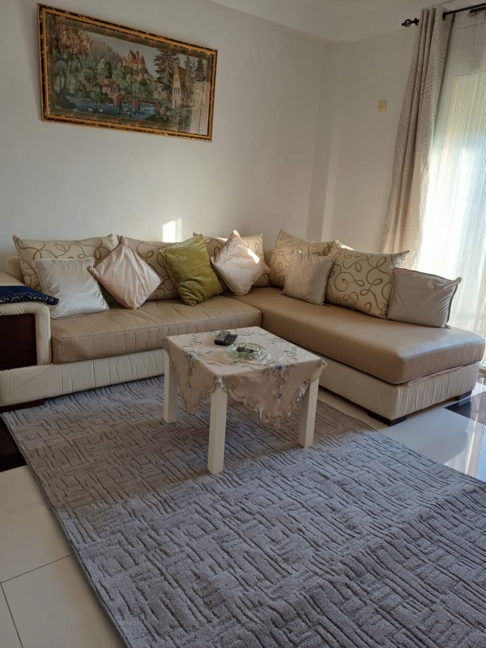 A comfortable seating area is presented with a stylish cream-colored sectional sofa adorned with multiple decorative cushions. A small, square coffee table rests atop a textured light grey rug, and a piece of wall art depicting a scenic landscape adds a touch of character to the space.