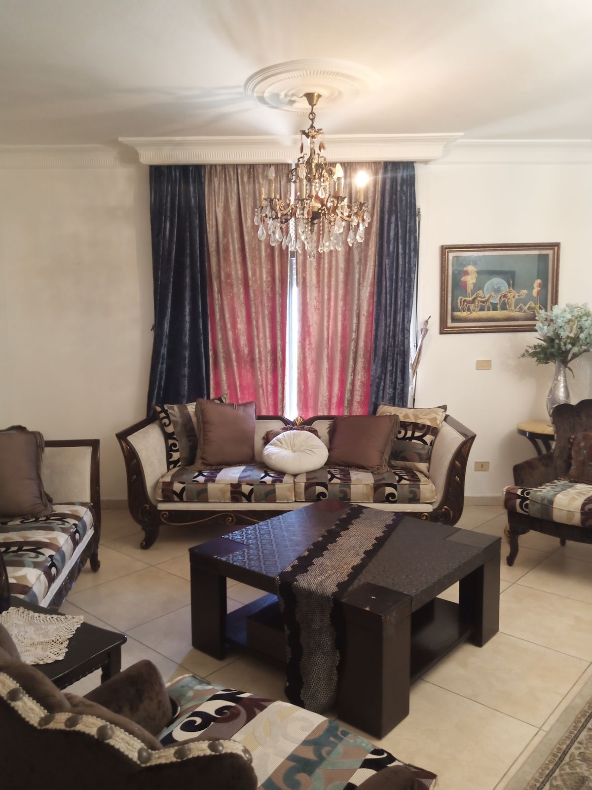 A well-furnished living area features intricately designed seating with a mix of dark wood and patterned upholstery. A decorative chandelier hangs from the ceiling above a stylish coffee table. Floor-to-ceiling curtains in rich colors frame a tasteful artwork on the wall.