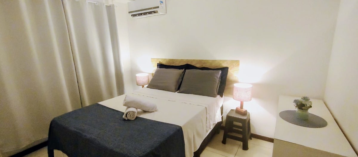 A well-appointed bedroom features a spacious bed with neutral bedding and accent pillows. Nightstands with soft lighting are positioned on either side, complementing the understated decor. A dresser is visible, and large curtains are drawn to the side, allowing light to fill the room.