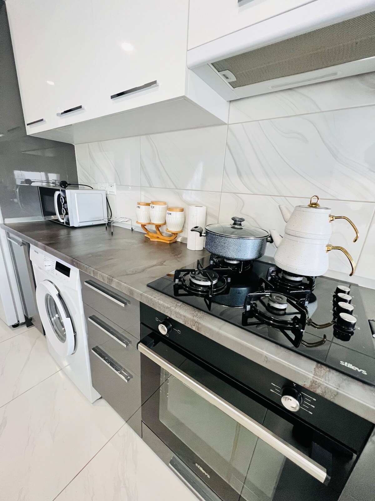 The kitchen is equipped with modern appliances, including a microwave, washing machine, and a sleek stovetop with two gas burners. Finished with a polished countertop, designed for both functionality and style, the space also features cookware and dining essentials neatly arranged.
