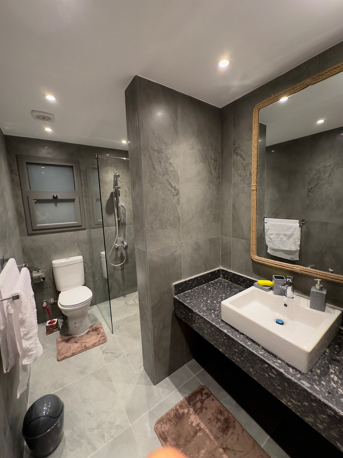 A modern bathroom is presented with sleek gray walls and marble tiles. A spacious shower enclosure with glass doors is visible alongside a toilet. A neatly arranged countertop features a rectangular sink and a large mirror framed in gold. Soft towels are hung near the sink area.