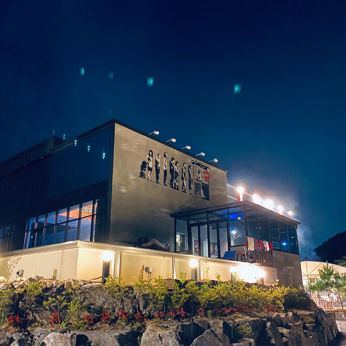 The exterior of a two-and-a-half-story house is illuminated against a night sky. A series of artistic paintings is displayed on the upper section of the building. Below, a well-manicured garden with colorful flowers and stone landscaping enhances the overall aesthetic.