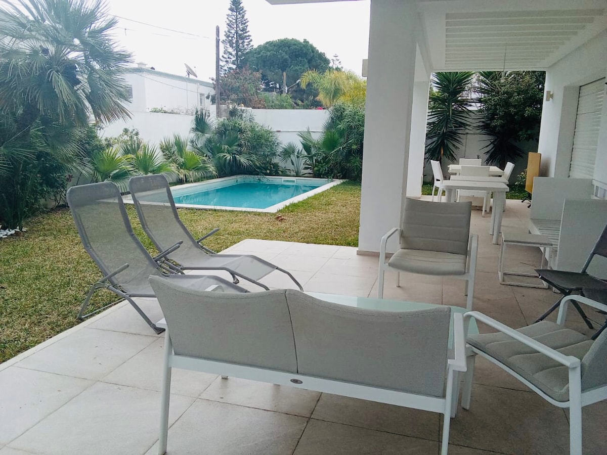 A serene outdoor area features a refreshing pool surrounded by lush greenery. Several comfortable lounge chairs and a dining table are positioned on a tiled patio, offering ample space for relaxation and enjoyment in a private garden setting.