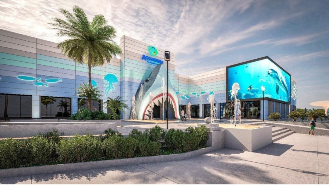 A modern aquarium building features an eye-catching façade with colorful aquatic murals and large glass windows revealing marine life displays. Fountains create a playful atmosphere in the landscaped entrance, while palm trees add to the tropical feel of the surroundings.