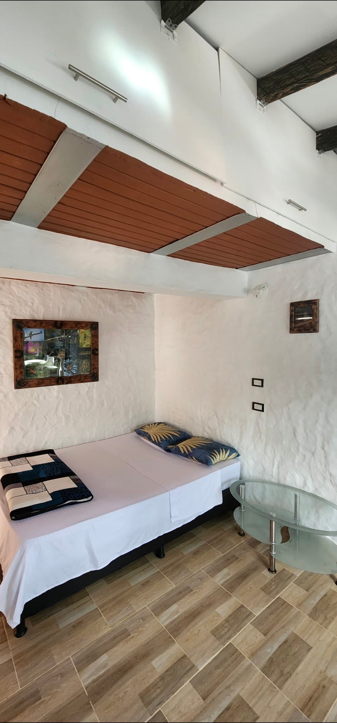 The room features a single bed dressed in white linens, complemented by patterned cushions. A small glass table is positioned nearby. Textured white walls enhance the cozy feel, and natural light fills the space through a window. A framed artwork adorns one wall.