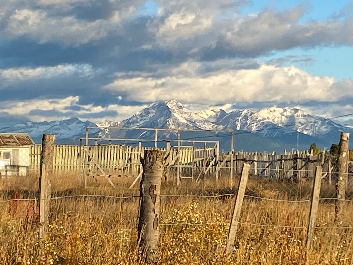 A scenic view features snow-capped mountains in the background, set against a blue sky with scattered clouds. In the foreground, a fenced area with wooden posts and dry grass is visible, creating a peaceful rural landscape.