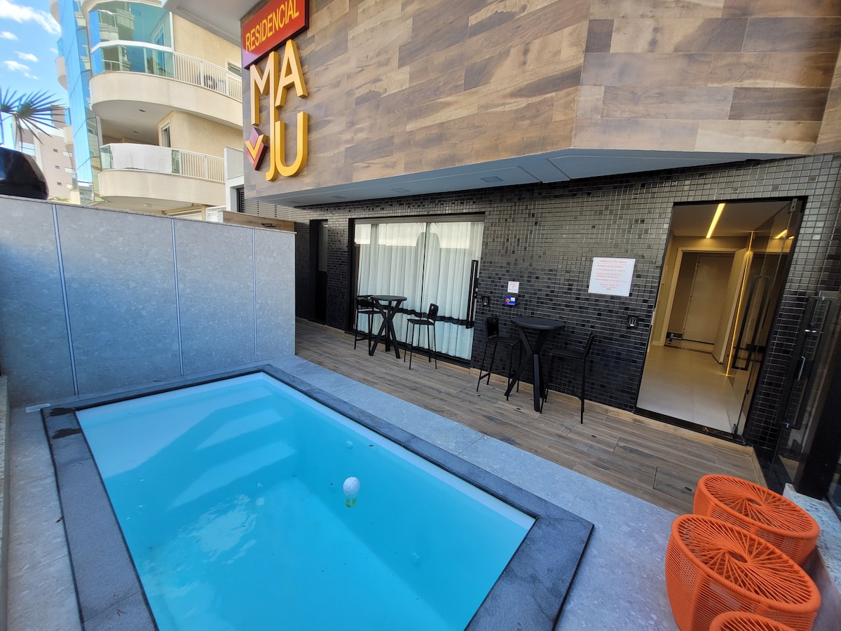 An outdoor area features a small swimming pool with clear water, framed by light stone tiles. Two black tables and four black chairs are positioned nearby. A wall of dark tiles and the building's signage are visible in the background.