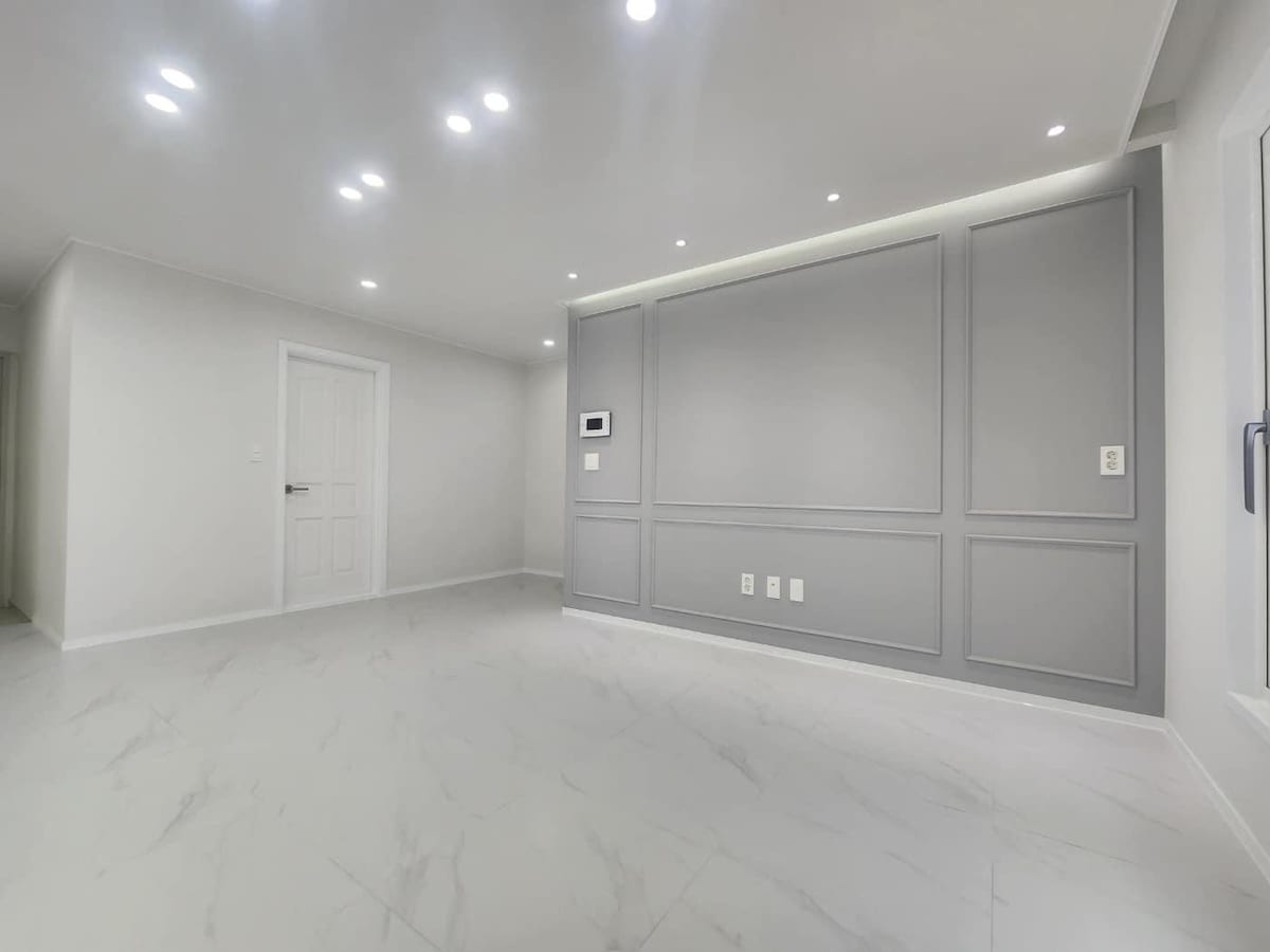 A spacious living area features a sleek, light-colored tiled floor and minimalist grey wall design. Recessed lighting illuminates the room evenly. A door leads to another space, while a control panel is discreetly mounted on the wall.