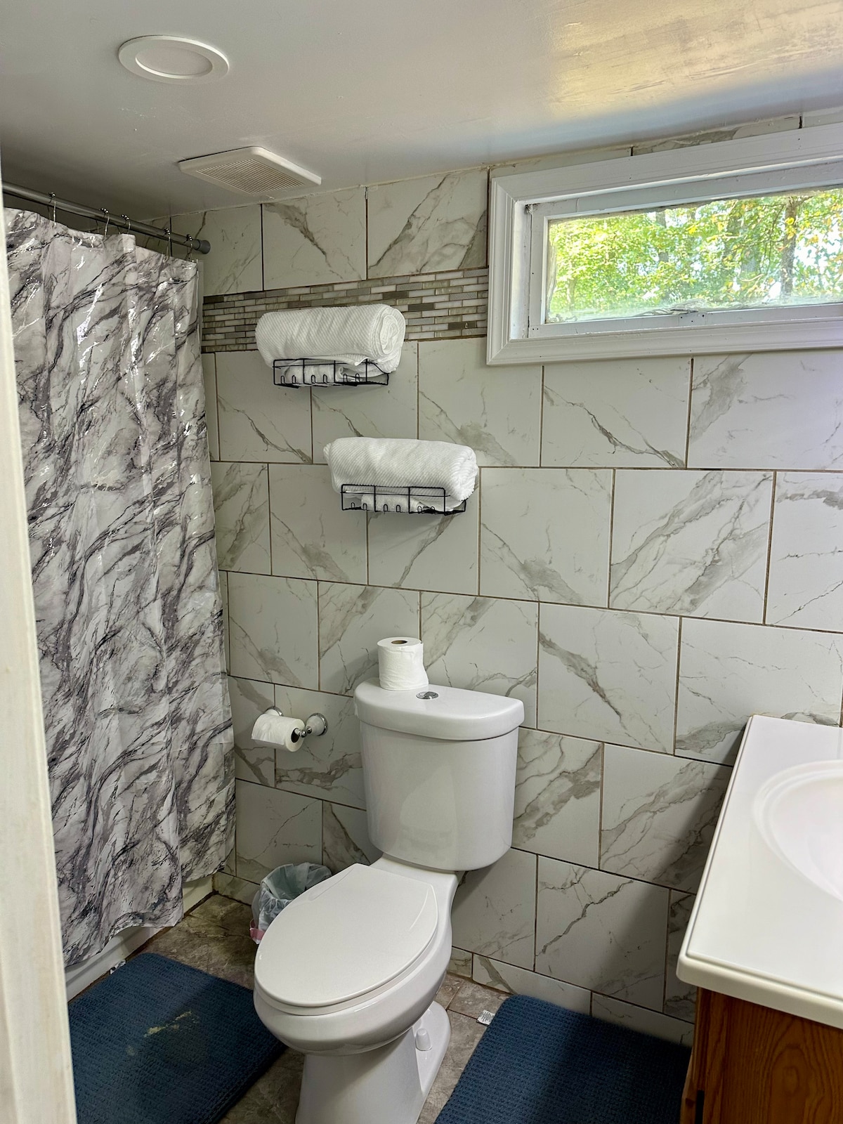The bathroom features a tiled wall design, with a shower area enclosed by a marble-patterned curtain. Two shelves hold neatly rolled towels, and a toilet paper roll is placed next to the toilet. Natural light flows in through a window above the sink.