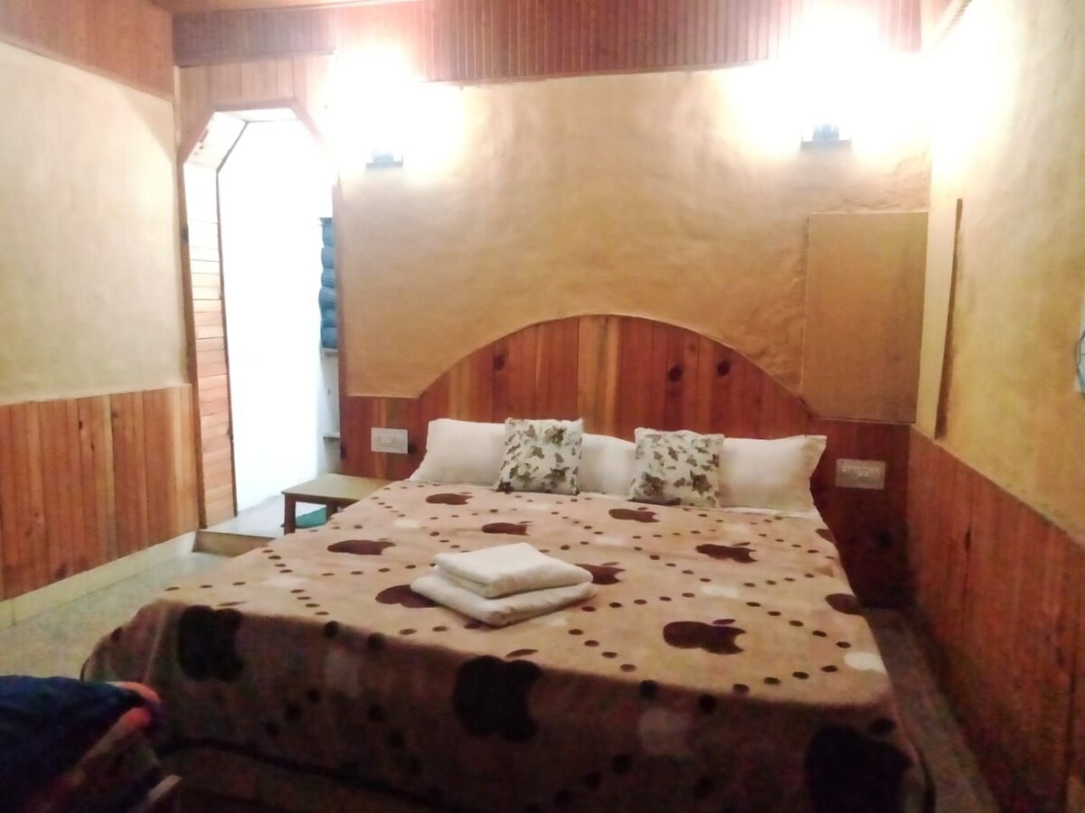 A cozy bedroom features warm wooden paneling and a spacious bed topped with a patterned blanket. Decorative pillows are arranged at the head of the bed. Soft lighting is provided by wall sconces, creating a comfortable atmosphere. A doorway leads to another space in the background.