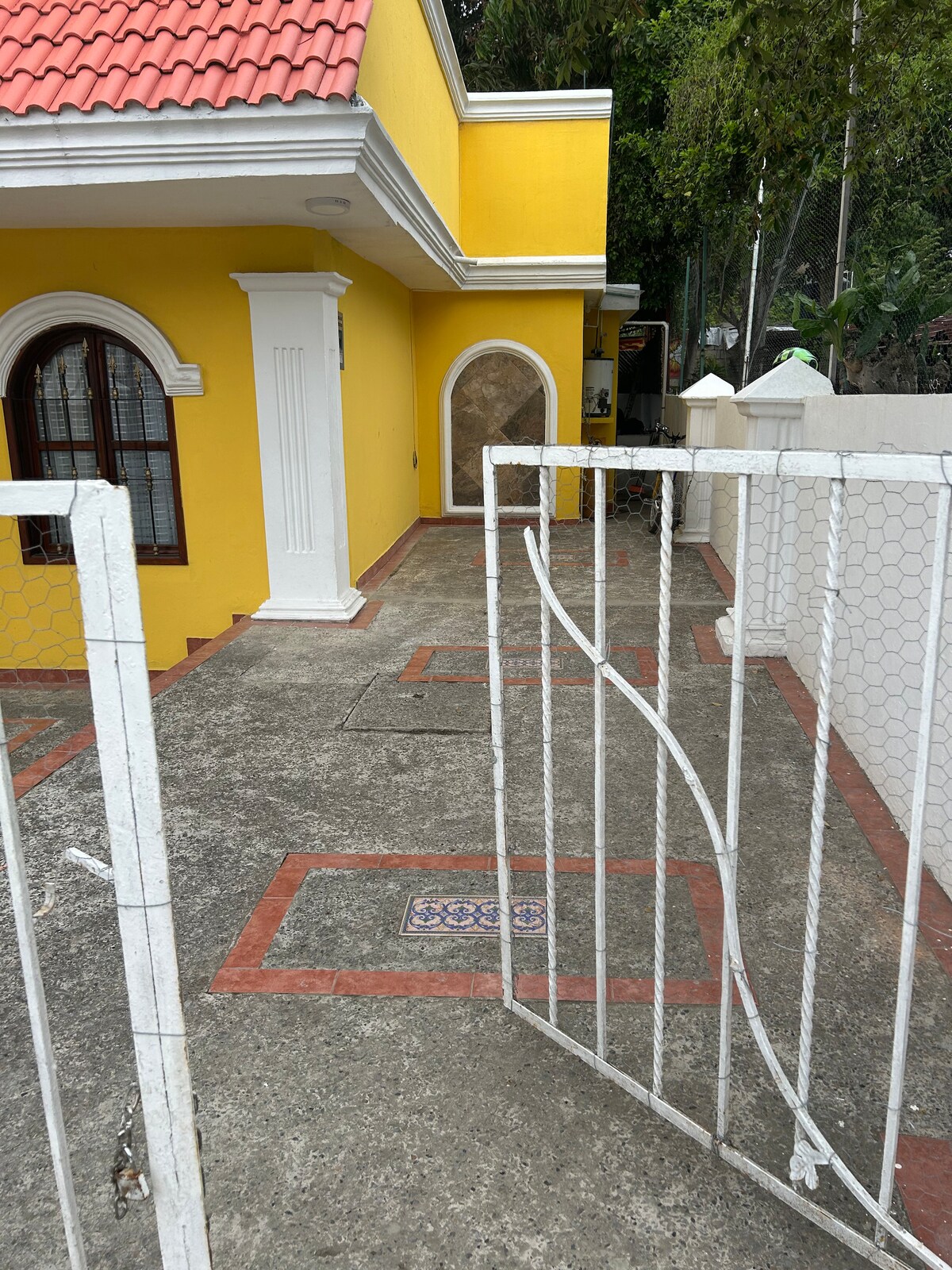 A vibrant yellow exterior of a house is seen, complemented by a red-tiled roof. A white gate, partially open, leads to a paved pathway. The entrance is framed by columns, and the surroundings feature greenery, adding a serene touch to the facade.