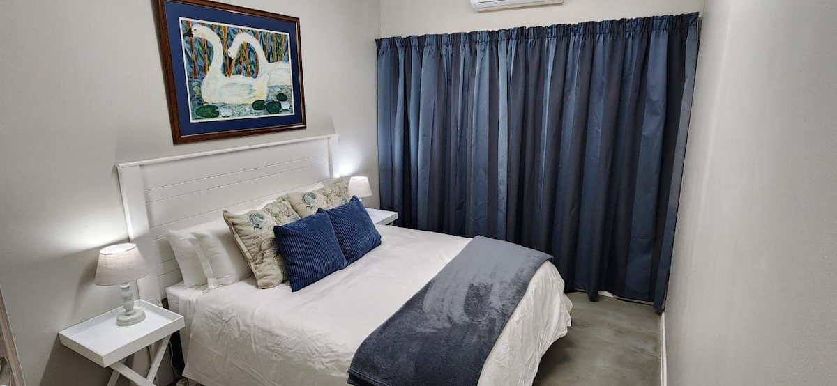 A well-lit sleeping area features a spacious bed dressed in white linens, accentuated by a dark throw blanket. Two bedside tables with lamps flank the bed, while blue drapes cover the window, offering privacy. A framed artwork is mounted on the wall above the headboard.