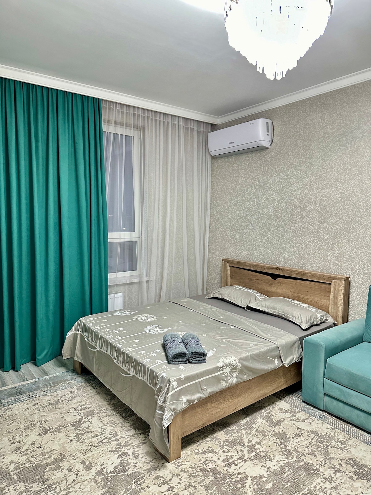 A serene bedroom features a wooden bed with a light-colored bedding set. Soft towels are neatly placed on the bed. Green curtains frame the window, and a cozy seating area is visible in a matching hue. A ceiling light adds brightness to the space.