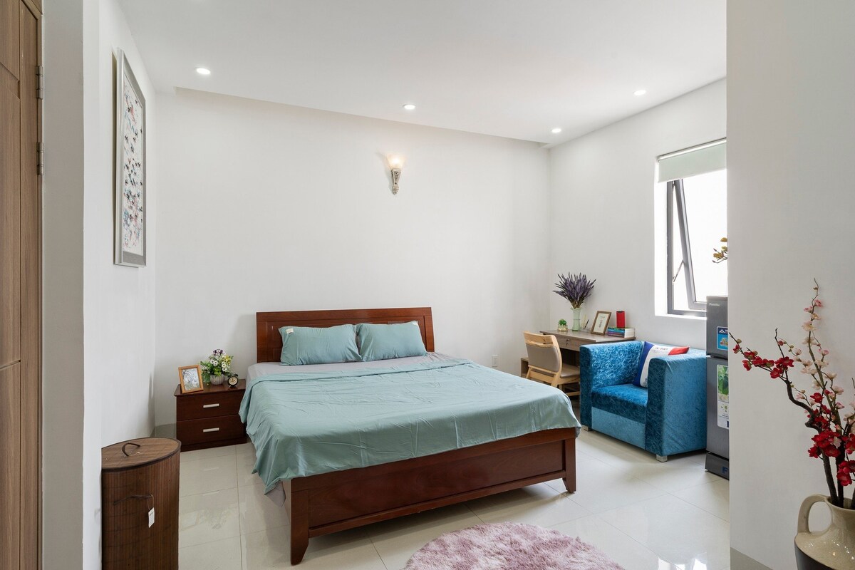 A bright and airy room features a spacious bed with a light blue cover. A wooden bedside table stands beside the bed, and a work desk with a chair is positioned near a window. A cozy blue chair adds to the inviting atmosphere.