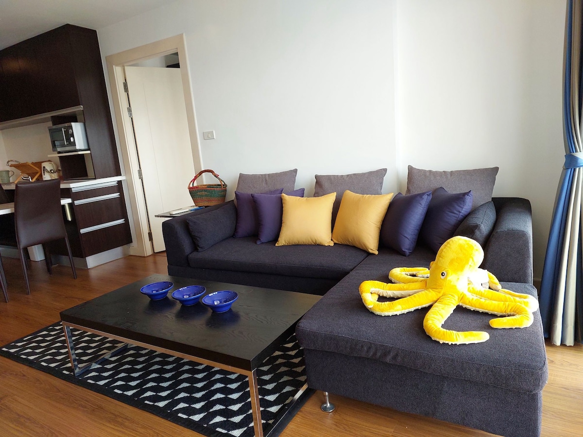 A modern living space is presented with a dark sofa adorned with yellow and purple cushions. A playful yellow octopus plush toy rests on the sectional. A sleek coffee table is positioned in front, accompanied by three blue bowls. The kitchen area and dining seating are visible in the background.