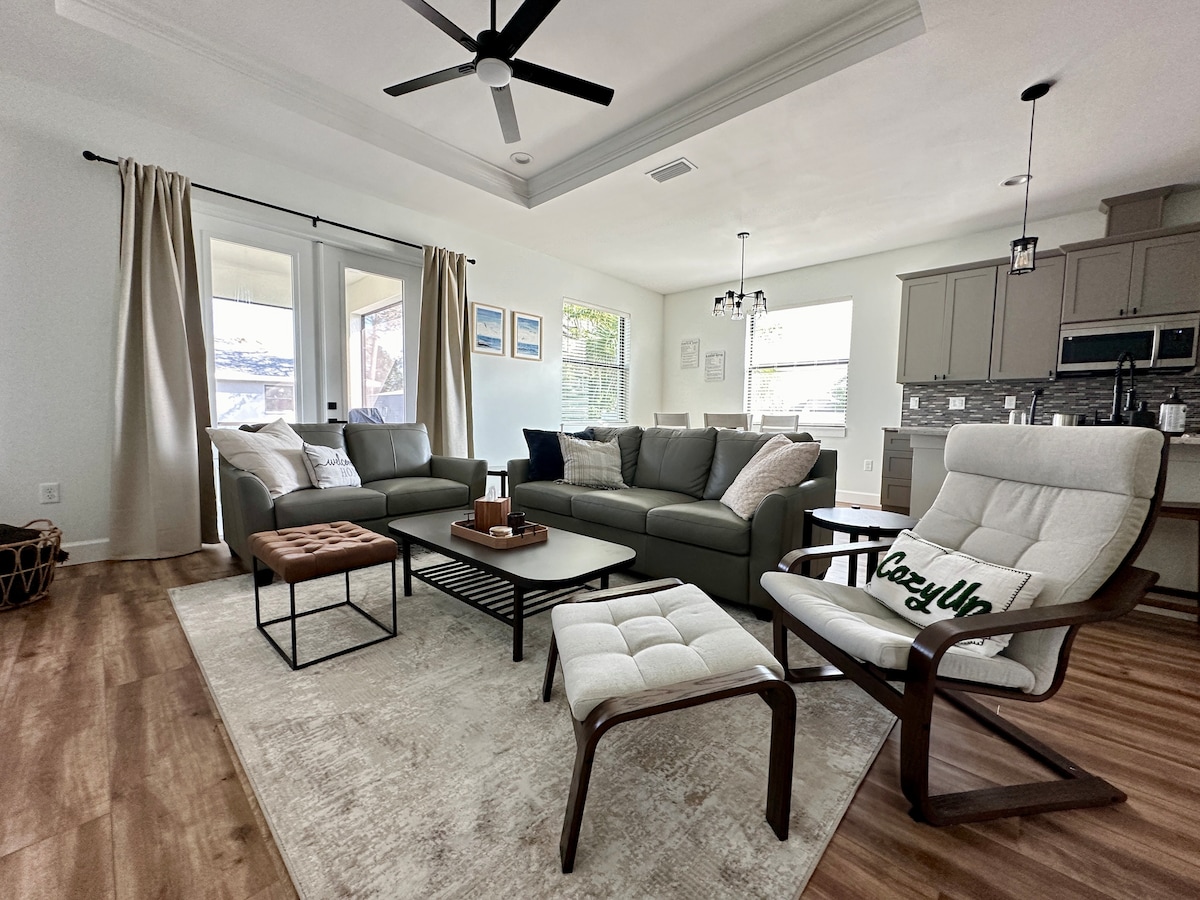 A spacious living area displays tall ceilings and an open layout. A large sectional sofa complements a coffee table, while a modern chair adds seating. Natural light floods in through windows, and light-colored curtains softly frame the space, enhancing the welcoming environment.