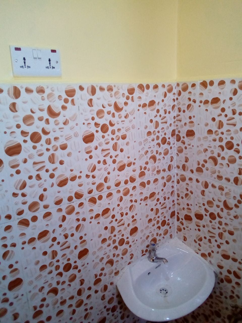 A small bathroom corner is displayed, featuring unique patterned wall tiles with a combination of earthy tones. A modern sink is located against the wall, complemented by a minimalist faucet. The walls are painted in a soft yellow, enhancing the space's brightness.