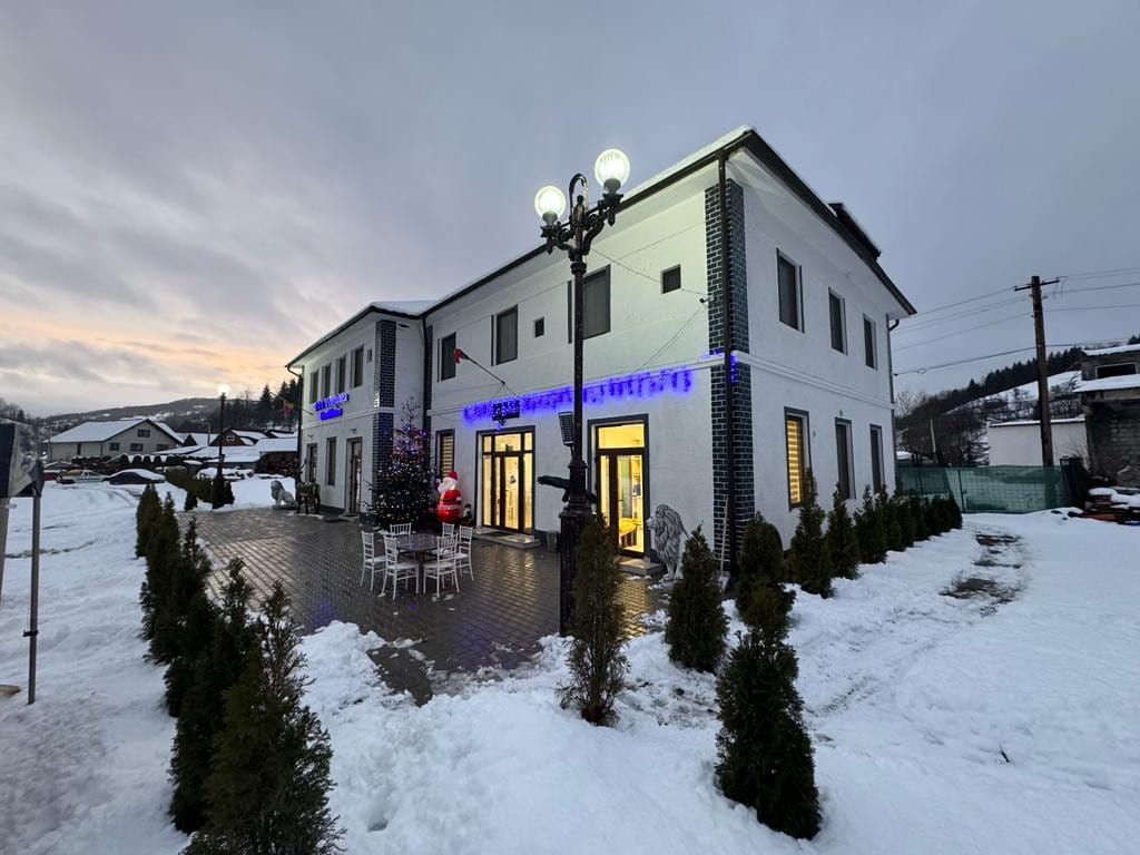 The exterior of the property is displayed, featuring a two-story building with light-colored walls and dark trim. Snow covers the ground, and small evergreen shrubs are arranged along the pathway. Outdoor seating with white chairs and a table is visible near the entrance, complemented by festive decorations.