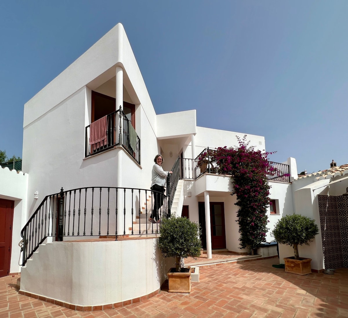The exterior of the house displays a bright, white facade with a distinctive balcony adorned by purple bougainvillea. A person can be seen approaching the entrance, which features a set of stairs and a wrought iron railing. The surrounding patio is complemented by two small trees in decorative pots.