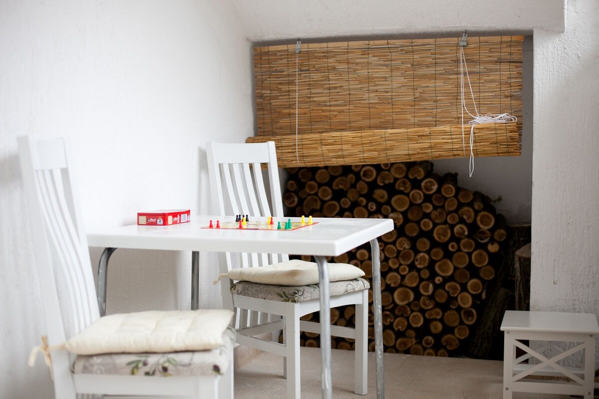 A small dining area features a white table accompanied by two white chairs with soft cushions. A stack of neatly arranged logs is visible in the background, while a bamboo shade partially covers the window, adding a natural touch to the space.
