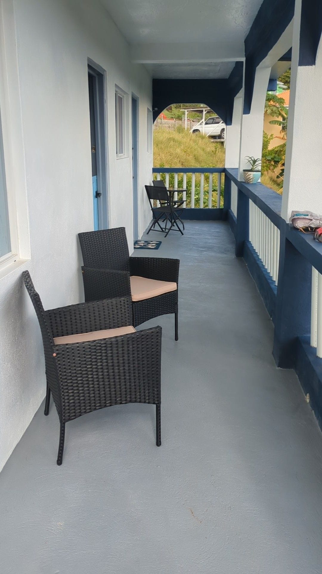 A shaded verandah is shown with two wicker chairs positioned on a smooth concrete surface. One chair is placed near the railing, while a smaller folding chair is visible in the background, offering a serene space for relaxation.