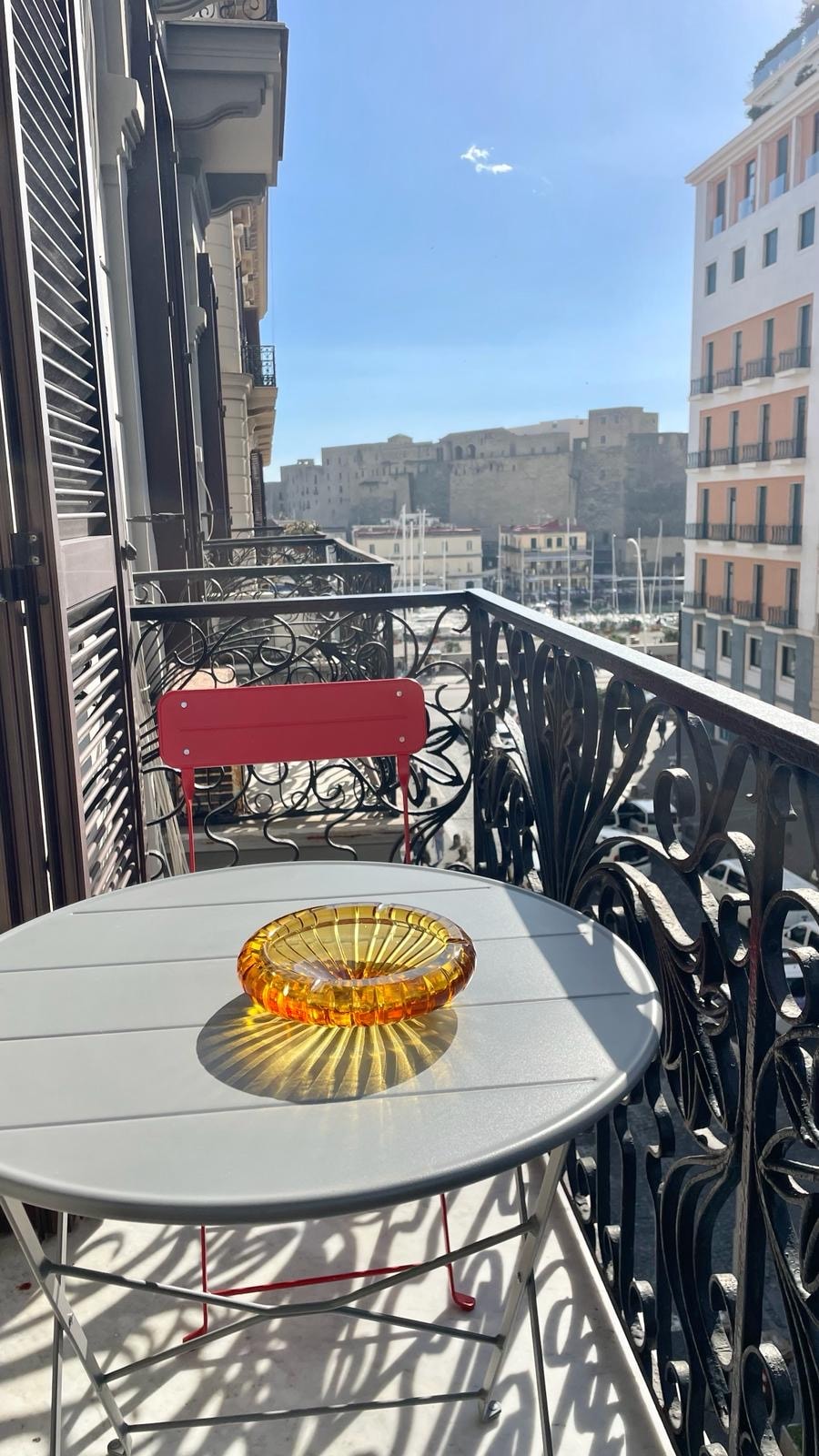 A small balcony is shown, featuring a round table and a red chair. An amber glass bowl rests on the table, reflecting sunlight. The ornate railing provides a view of the surrounding buildings and the harbor.