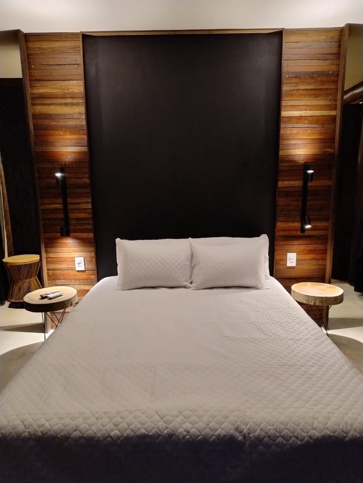 A spacious bed is positioned against a dark accent wall, flanked by two wooden side tables. Soft lighting from wall sconces creates a calming ambiance, while the textured wood paneling adds warmth to the room.