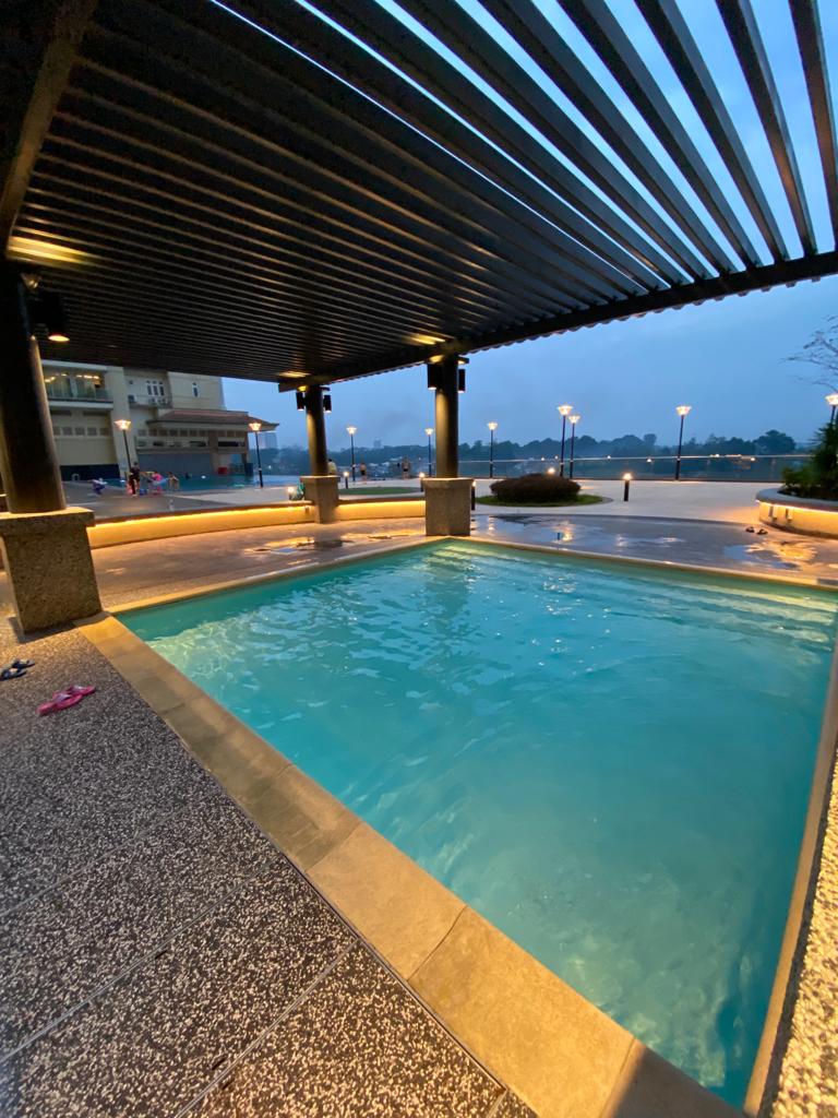 A private swimming pool is showcased beneath a slatted pergola, featuring clear blue water. Surrounding lights create a subtle ambiance, reflecting off the pool's surface. The distant view includes a building and pathways along the water's edge, enhancing the serene environment.
