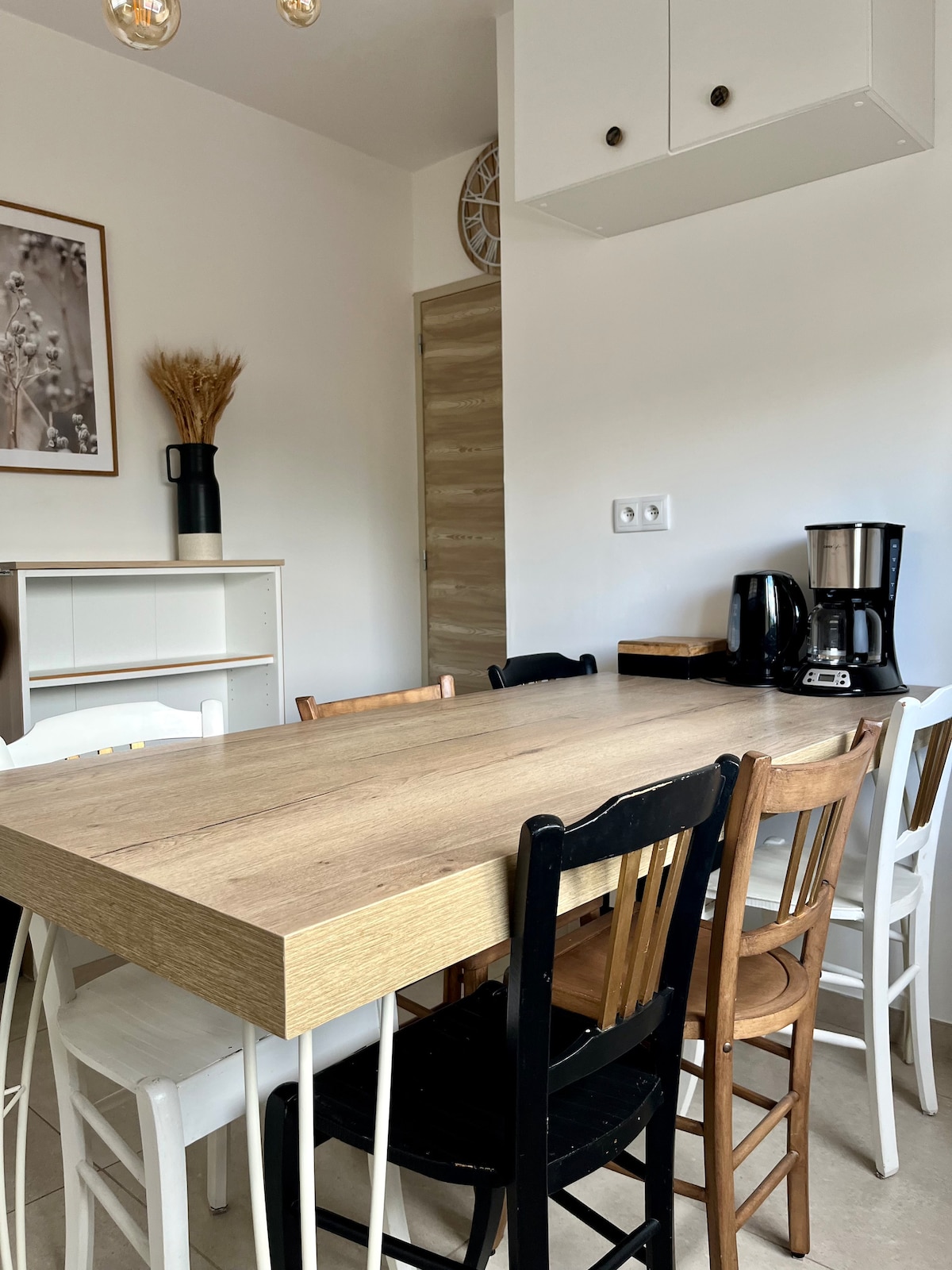 A wooden dining table is surrounded by a mix of eight chairs in different colors, creating an inviting dining area. A coffee maker and a kettle are placed next to a small cabinet, with a minimalist artwork visible on the wall.