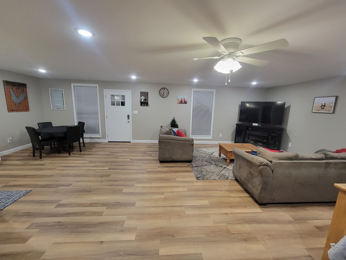 A spacious living area features comfortable seating arrangements with a light-colored couch and matching loveseat. A dining table is set for four, while large windows allow natural light to fill the room. A TV is positioned against a wall, complemented by soft overhead lighting.