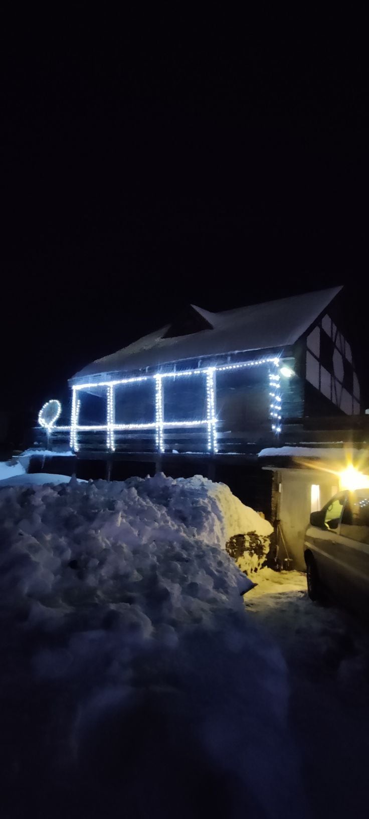A house exterior is adorned with white string lights, creating a festive ambiance against a backdrop of dark night sky. Snow piles accumulate around the base of the house, reflecting warm light from a nearby vehicle parked on the side.