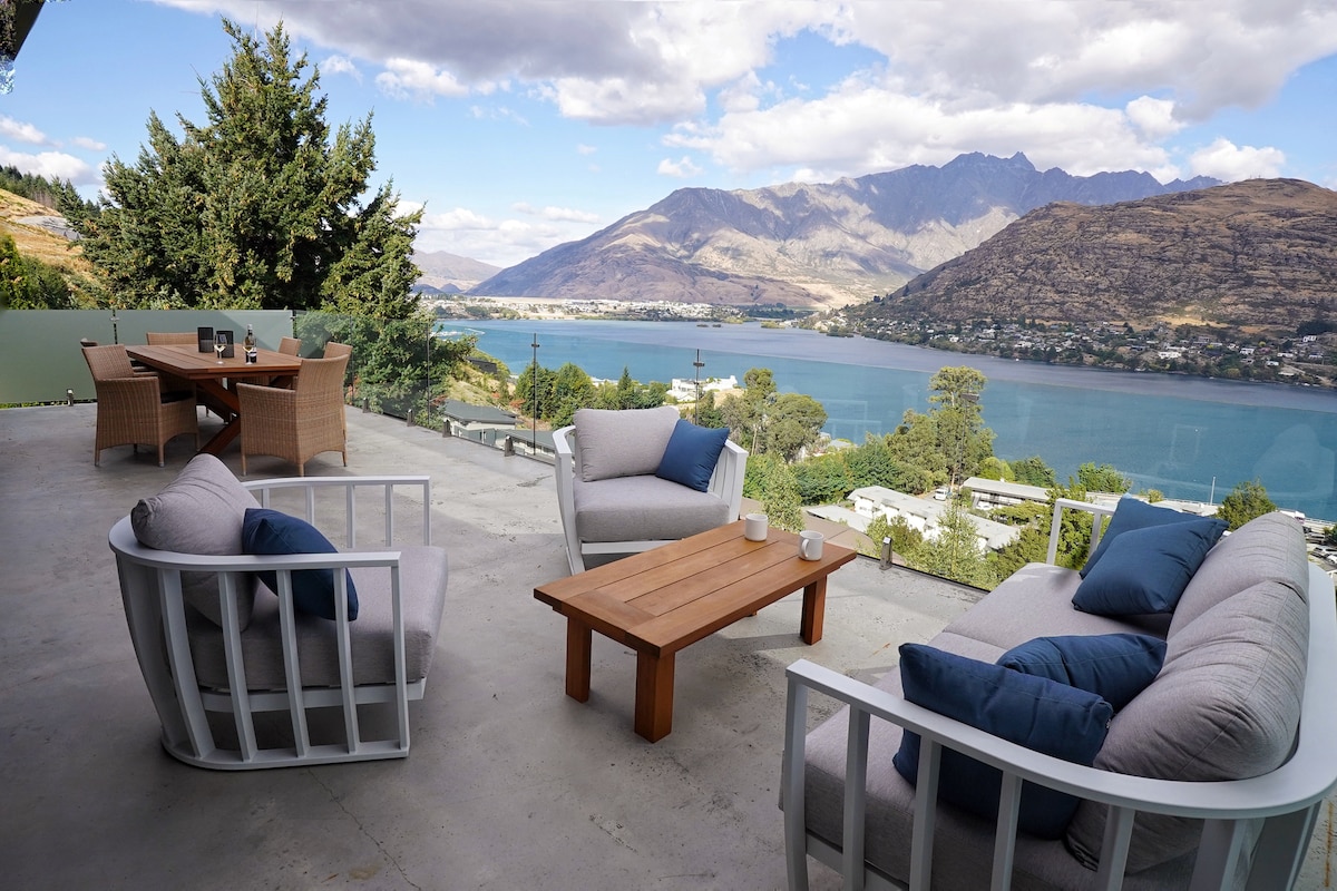 An outdoor seating area features cozy lounge chairs and a wooden table, positioned to take in views of Lake Wakatipu and the Remarkable Mountain range. Tall trees surround the space, offering a sense of tranquility amidst the scenic landscape.