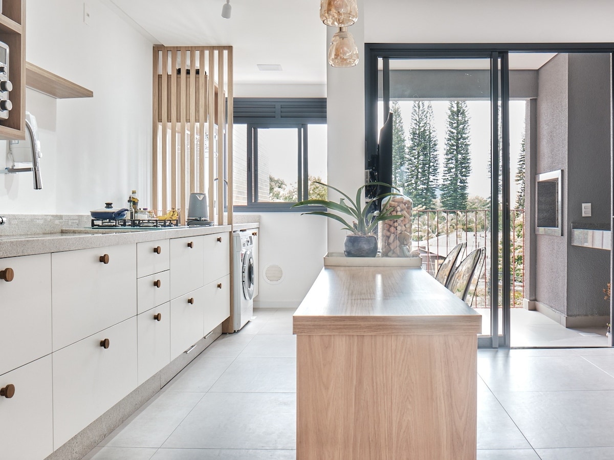 A modern kitchen is showcased, featuring sleek cabinetry and a spacious island. Natural light filters in through large sliding doors, revealing a view of outdoor greenery. A washing machine is positioned beside the cabinetry, enhancing the kitchen's functionality.