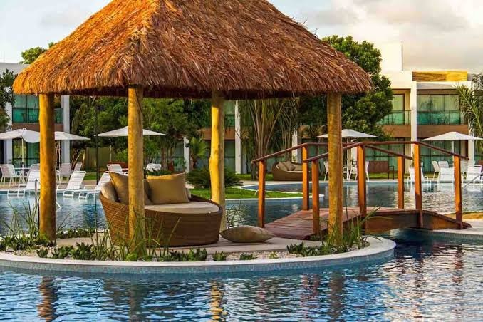A thatched-roof gazebo is situated on an island surrounded by water. A wooden bridge connects the gazebo to a larger pool area, which features several lounge chairs and umbrellas lining the water's edge. Lush greenery encircles the pool.