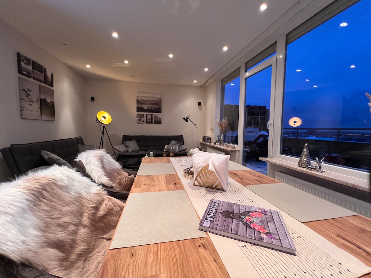 The bright living and dining area showcases large windows with a view of the outside. A wooden dining table is set with neutral table runners and decorated with a magazine. Two dark sofas provide ample seating, complemented by stylish lamps and wall decor.
