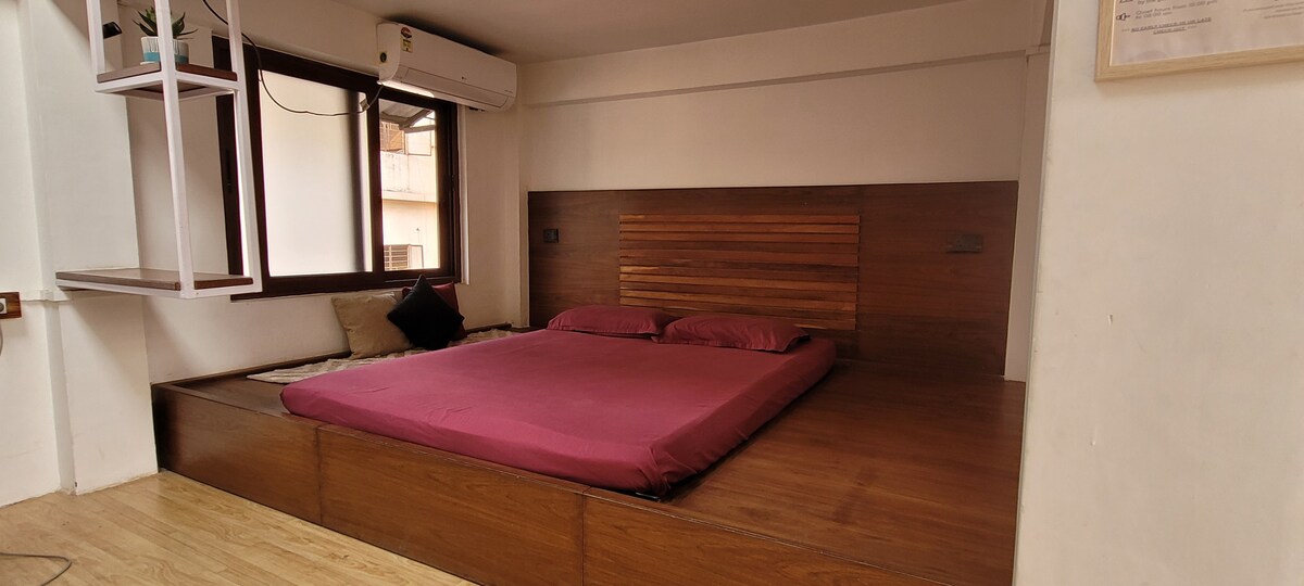 The space features a platform bed framed with rich wood tones, complemented by soft, burgundy bedding. A window provides natural light, while a cozy nook with cushions is situated nearby, enhancing the room's overall comfort and functionality.