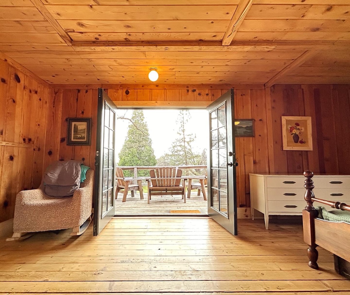 An interior view of the cabin showcases rustic wooden walls and floors. Two French doors open to a spacious deck with wooden chairs. A cozy seating area is visible, featuring a textured chair and a white dresser, creating a welcoming atmosphere.