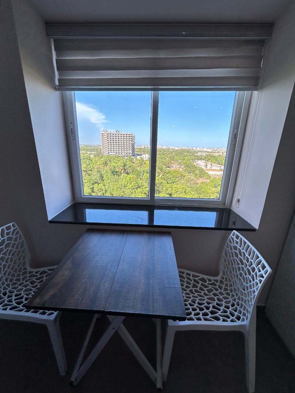A window offers a view of lush greenery and distant buildings. A small, minimalist table is positioned beside the window, accompanied by two white chairs featuring an intricate design. Natural light enters the space, highlighting a modern and open atmosphere.