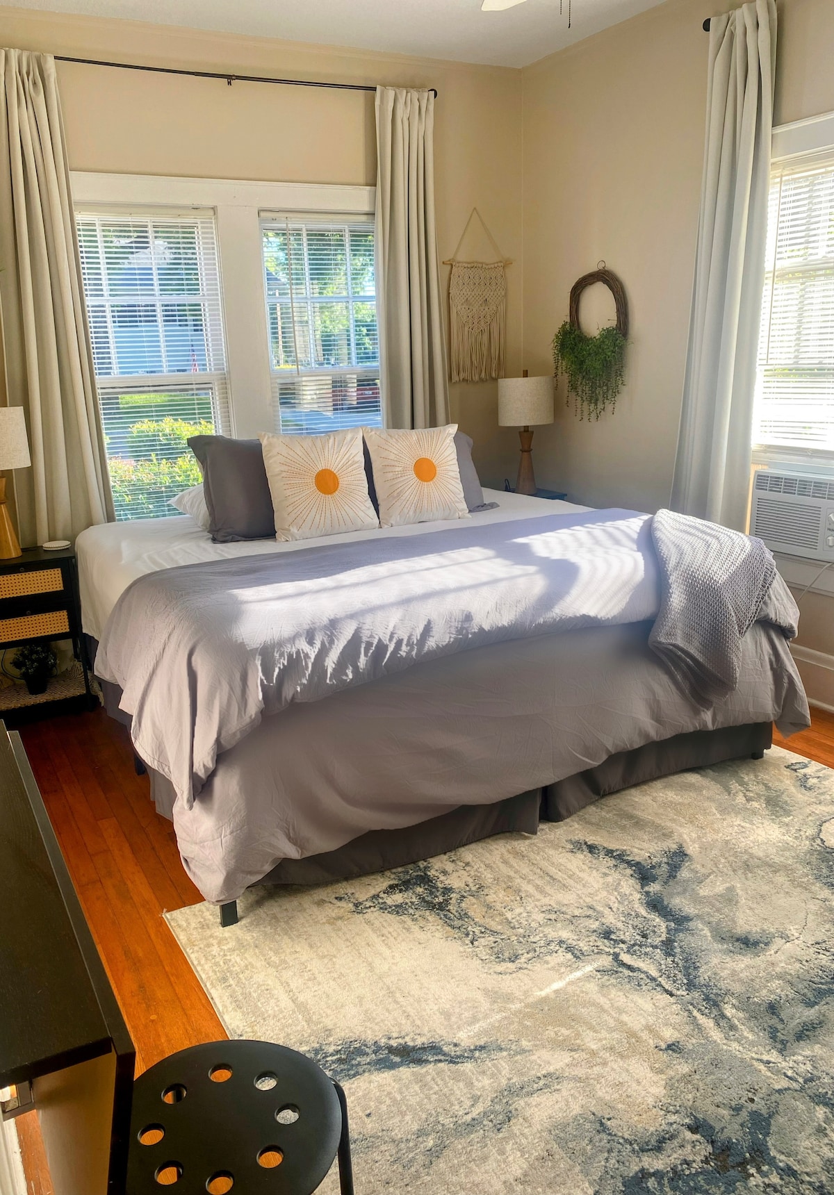 A spacious private room is highlighted by a large bed with muted bedding and decorative pillows featuring sun patterns. Natural light filters through multiple windows, creating a bright atmosphere. A tasteful area rug complements the wooden floor, and a small black stool adds a functional touch.
