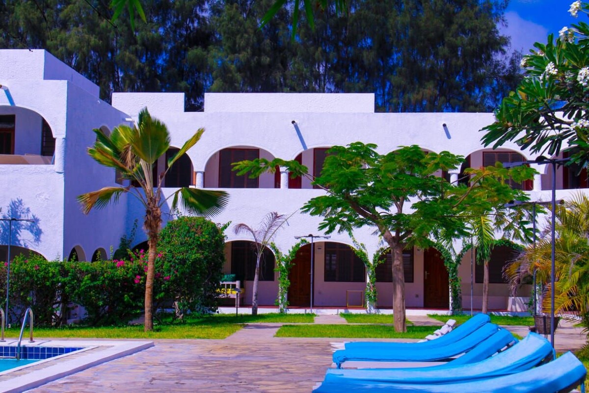The image showcases a modern two-story building with a white exterior and arched doorways. Lush green landscaping and palm trees frame the scene, while lounge chairs in blue are positioned near a pool, inviting relaxation in the serene outdoor environment.