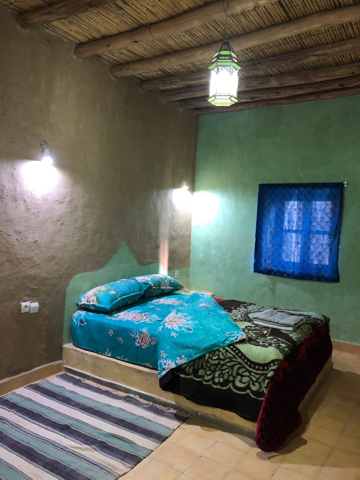 A cozy sleeping area features a spacious bed with a colorful turquoise and patterned blanket. The walls are accented in earthy tones, illuminated by warm wall lights. A small window with blue curtains allows natural light to enter, complementing the woven rug on the floor.