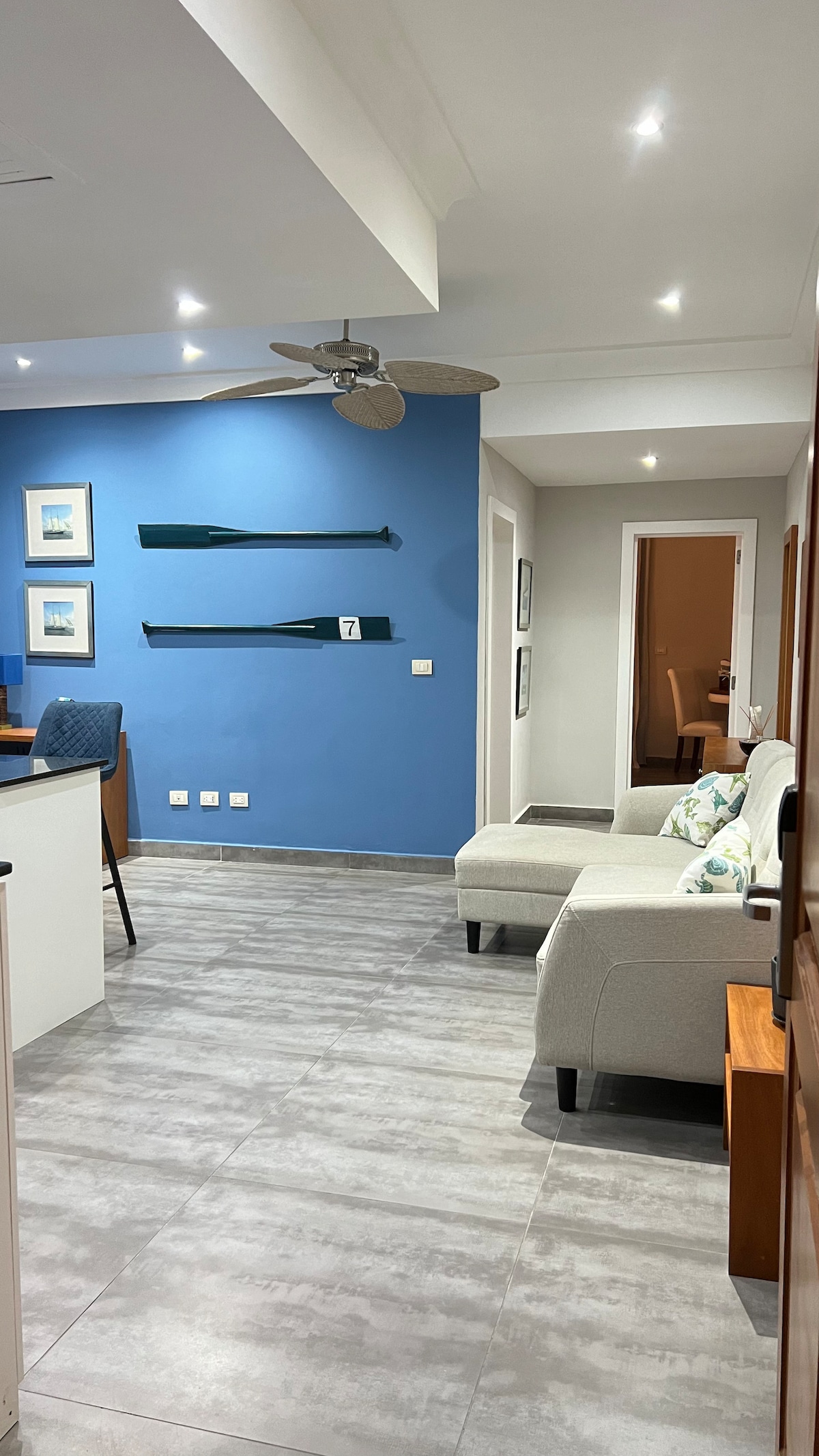 A well-lit living area features light grey furniture with decorative pillows. A blue accent wall enhances the space, while wooden oars are displayed decoratively. The room is completed with modern lighting fixtures and a view into an adjoining room.